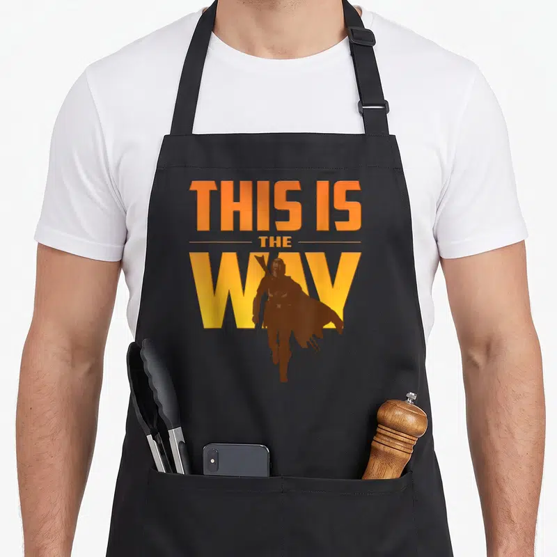 Tablier de cuisine noir avec texte This Is The Way et silhouette du Mandalorian en orange et jaune.