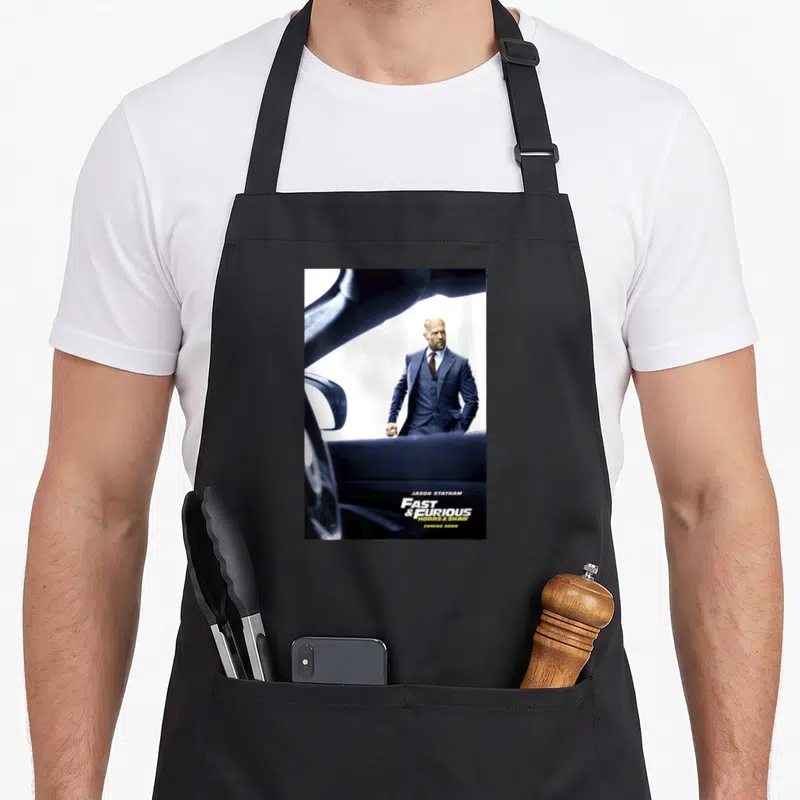 Tablier de cuisine noir avec une illustration de Jason Statham en costume bleu près d'une voiture.