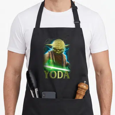 Tablier de cuisine noir avec une illustration de Yoda tenant un sabre laser vert sous son nom en lettres capitales dorées.