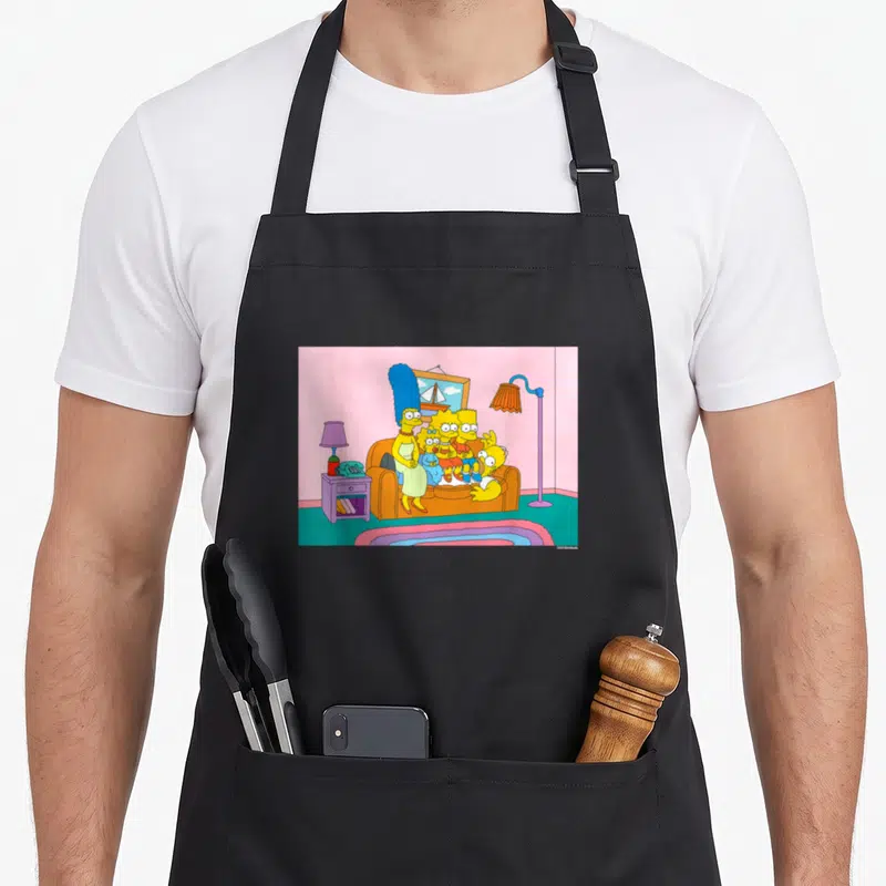 Tablier de cuisine noir avec illustration des Simpson sur leur canapé dans un salon rose et turquoise.