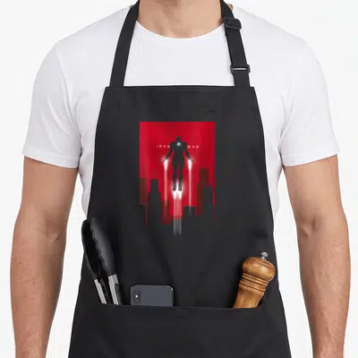 Tablier de cuisine noir avec une illustration d'Iron Man en vol sur fond rouge et silhouettes de bâtiments gris.