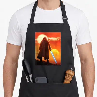 Tablier de cuisine noir avec une illustration d'Obi-Wan Kenobi tenant un sabre laser devant deux soleils orange.