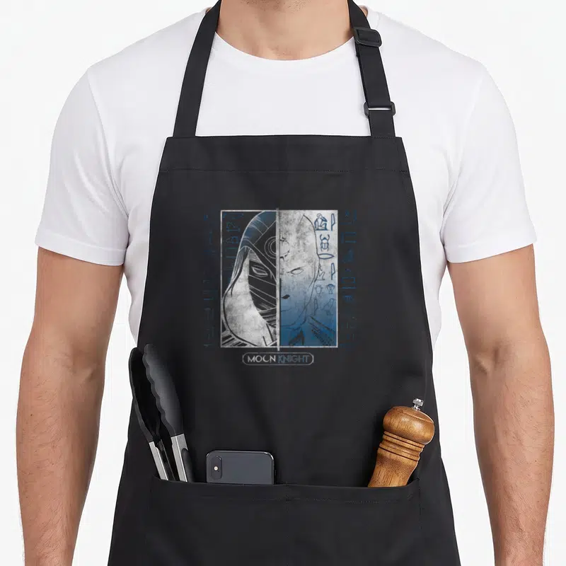 Tablier de cuisine noir avec illustration Moon Knight Split Face et hieroglyphes bleus sur fond texturé blanc.