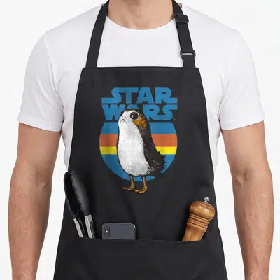 Tablier de cuisine noir avec illustration d'un Porg blanc et noir devant un logo Star Wars Last Jedi bleu vintage.
