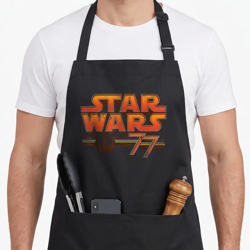 Tablier de cuisine noir avec logo Star Wars 1977 et symboles rétro en orange et brun.