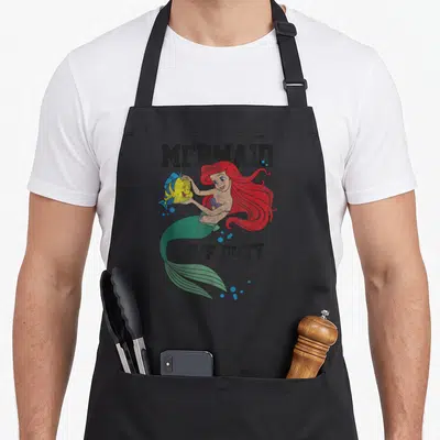 Tablier de cuisine noir avec Ariel et Flounder illustrés en style vintage sous le texte MERMAID OFF DUTY.