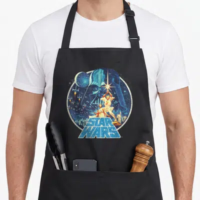 Tablier de cuisine noir avec illustration circulaire rétro Star Wars montrant Darth Vader et des vaisseaux spatiaux.