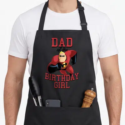 Tablier de cuisine noir avec illustration d'un super-héros en costume rouge et texte Dad of the anniversaire Girl en lettres capitales rouges.