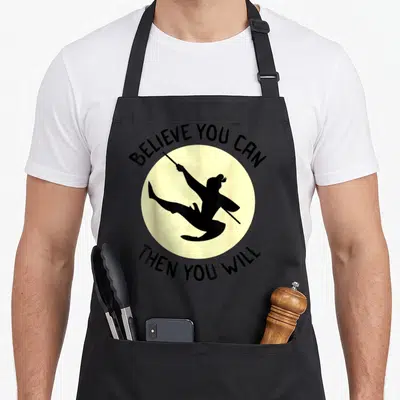 Tablier de cuisine noir avec silhouette Mulan et texte Believe You Can Then You Will sur fond circulaire jaune pâle.