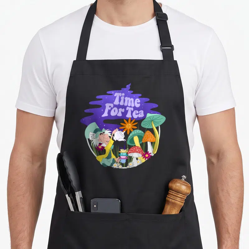 Tablier de cuisine noir avec illustration colorée du Mad Hatter versant le thé entre des champignons et des fleurs dans un décor violet.