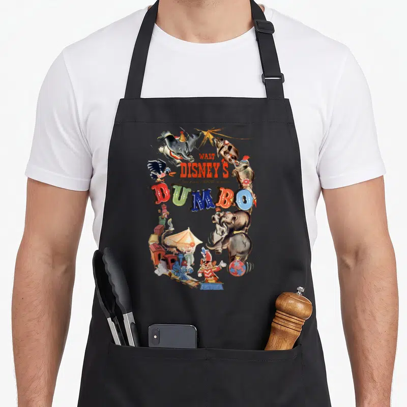Tablier de cuisine noir avec une illustration colorée de Dumbo et des personnages du cirque sur fond d'affiche de film vintage.