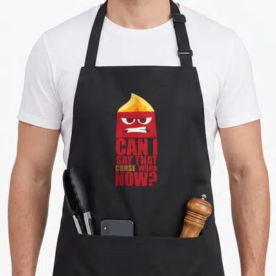 Tablier de cuisine noir avec personnage rouge à tête de feu et texte Can I Say That Curse Word Now en lettres capitales.