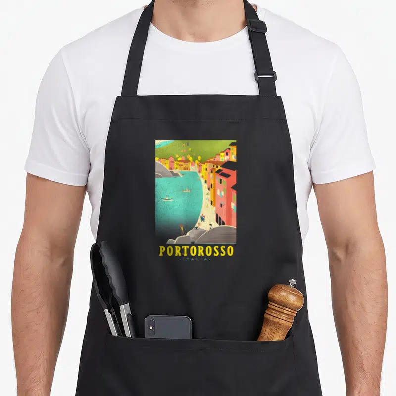 Tablier de cuisine noir avec une illustration colorée représentant le village italien de Portorosso surplombant la mer turquoise.