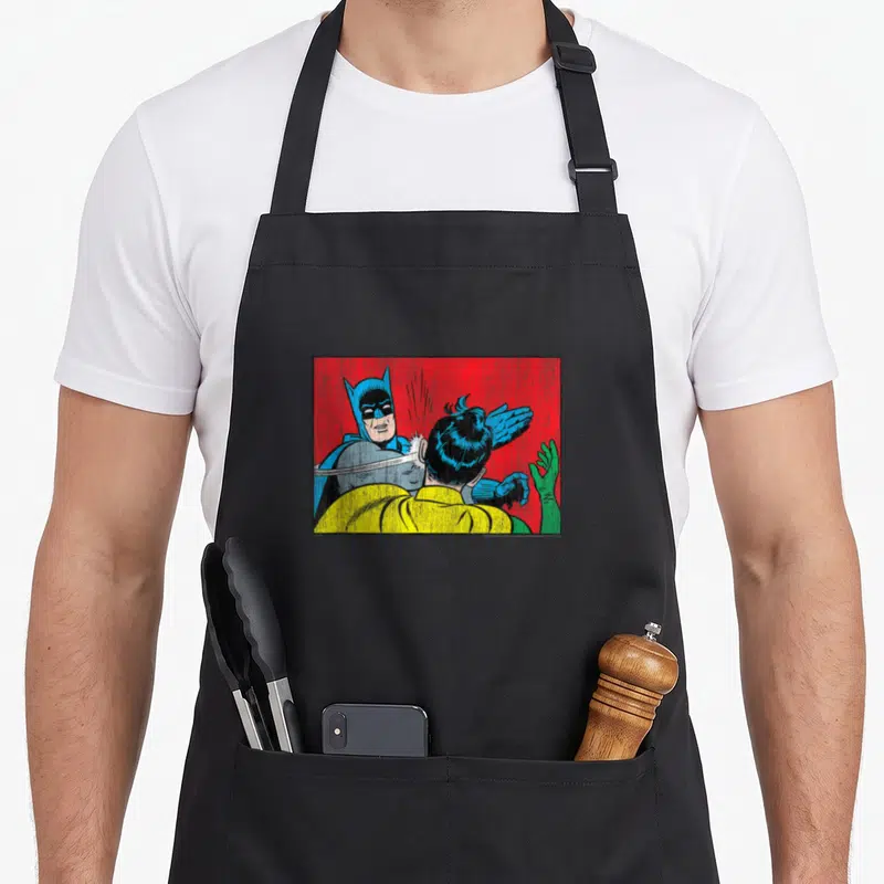 Tablier de cuisine noir avec une illustration colorée de Batman donnant une tape à Robin sur fond rouge vif.