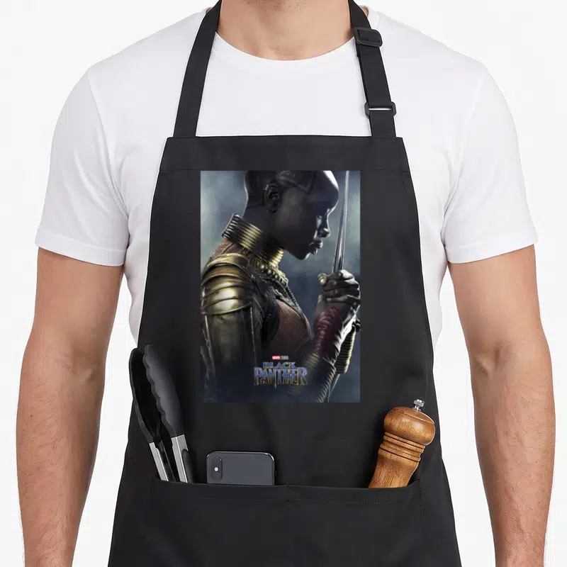 Tablier de cuisine noir avec un portrait latéral d'Okoye tenant une lance dorée et rouge, logo Black Panther en bas.
