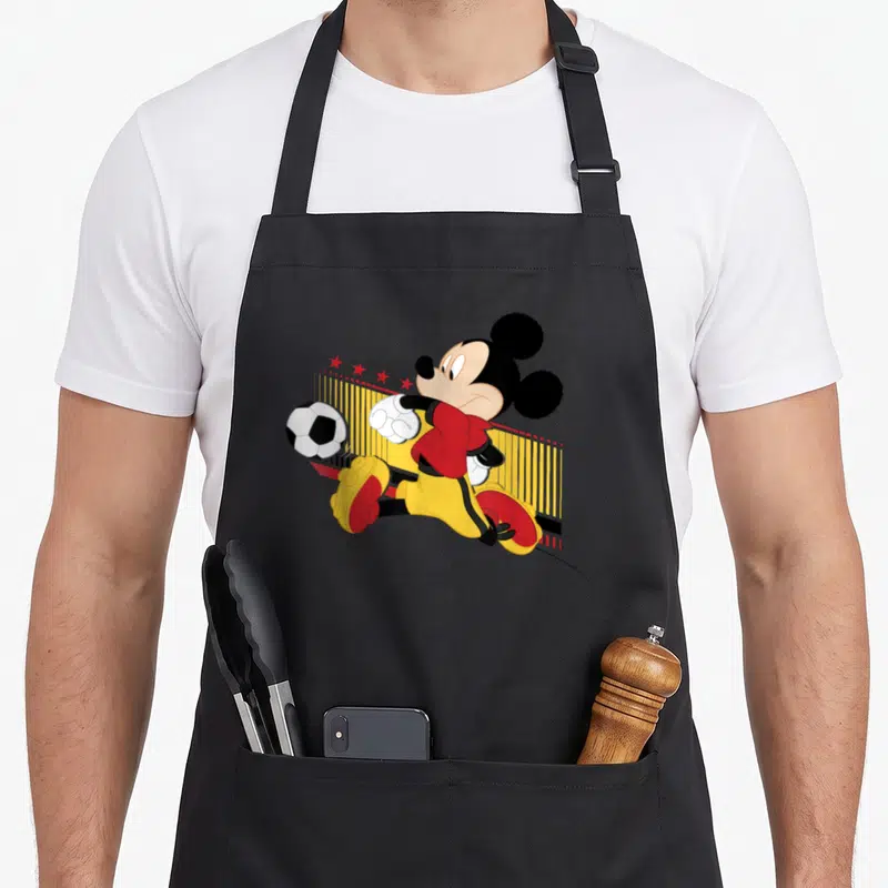Tablier de cuisine noir avec Mickey Mouse en maillot de football jaune et rouge frappant une balle.