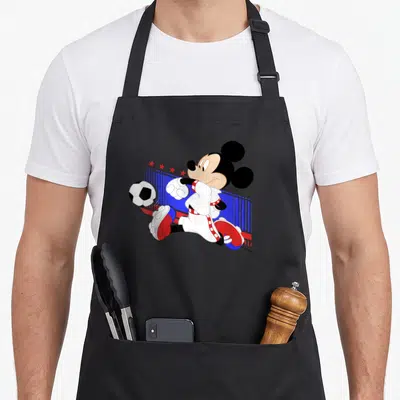 Tablier de cuisine noir avec Mickey Mouse en maillot blanc tirant sur un ballon de football devant des barres bleues et des étoiles rouges.