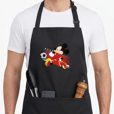 Tablier de cuisine noir avec Mickey Mouse en maillot rouge jouant au soccer devant un filet.