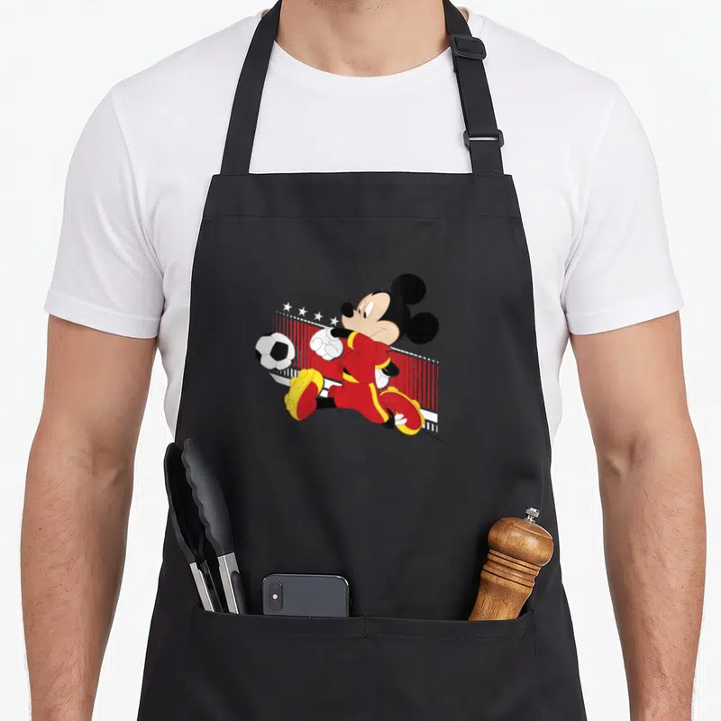 Tablier de cuisine noir avec Mickey Mouse en maillot rouge jouant au soccer devant un filet.