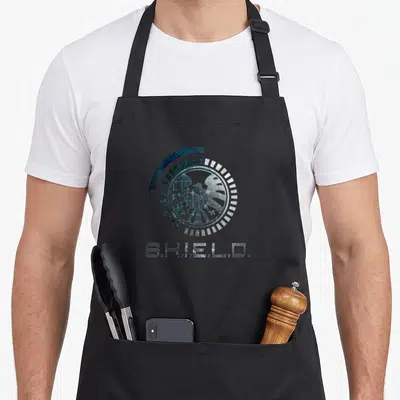 Tablier de cuisine noir avec le logo circulaire S.H.I.E.L.D. et un aigle au centre sur fond gris.