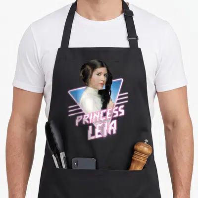 Tablier de cuisine noir avec une illustration de Princess Leia tenant un blaster devant un triangle bleu et le texte PRINCESS LEIA en rose.