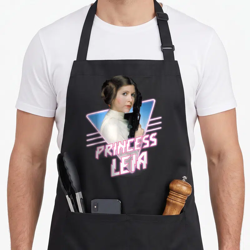 Tablier de cuisine noir avec une illustration de Princess Leia tenant un blaster devant un triangle bleu et le texte PRINCESS LEIA en rose.