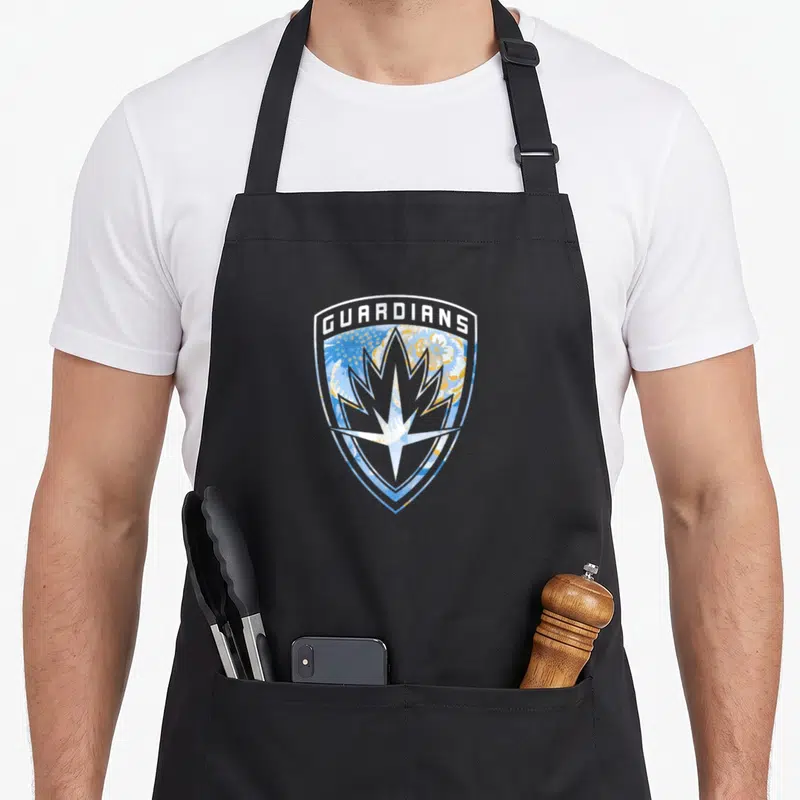 Tablier de cuisine noir avec un insigne en forme de bouclier orné de fleurs et d'une étoile centrale.