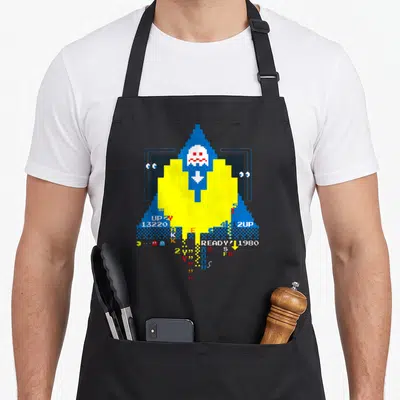 Tablier de cuisine noir avec un motif pixel art jaune et bleu représentant un fantôme et des éléments de jeu vidéo rétro.