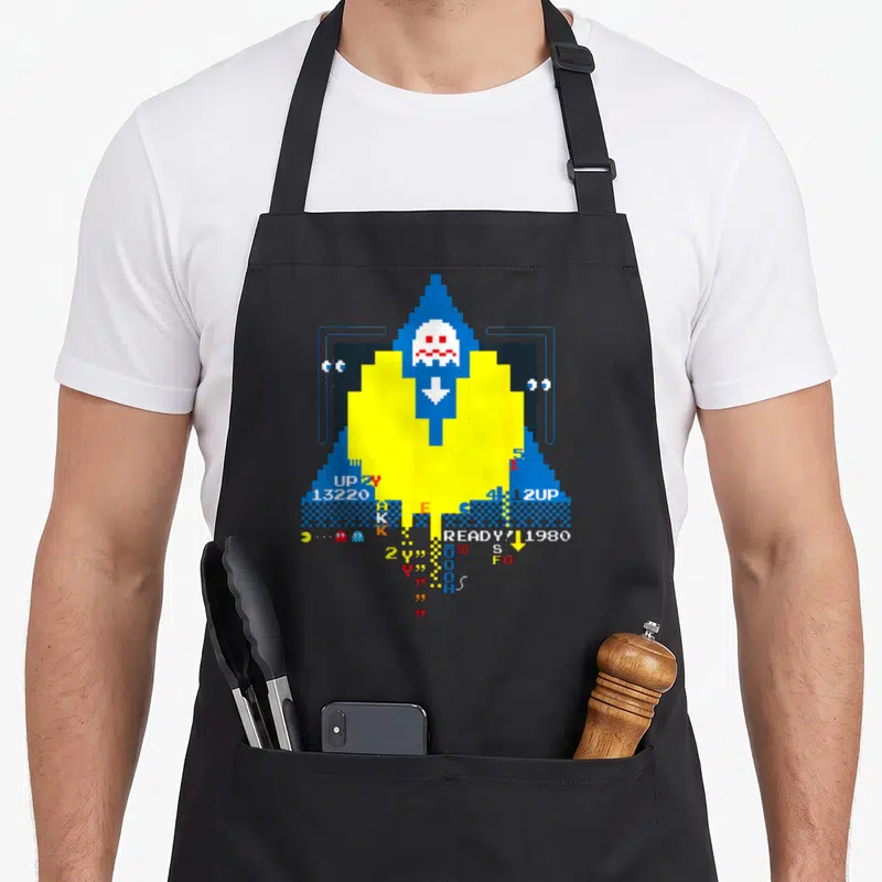 Tablier de cuisine noir avec un motif pixel art jaune et bleu représentant un fantôme et des éléments de jeu vidéo rétro.