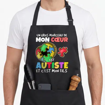 Tablier de cuisine noir avec un cœur en puzzle multicolore et un texte affirmant l'autisme du fils imprimé en lettres vives.