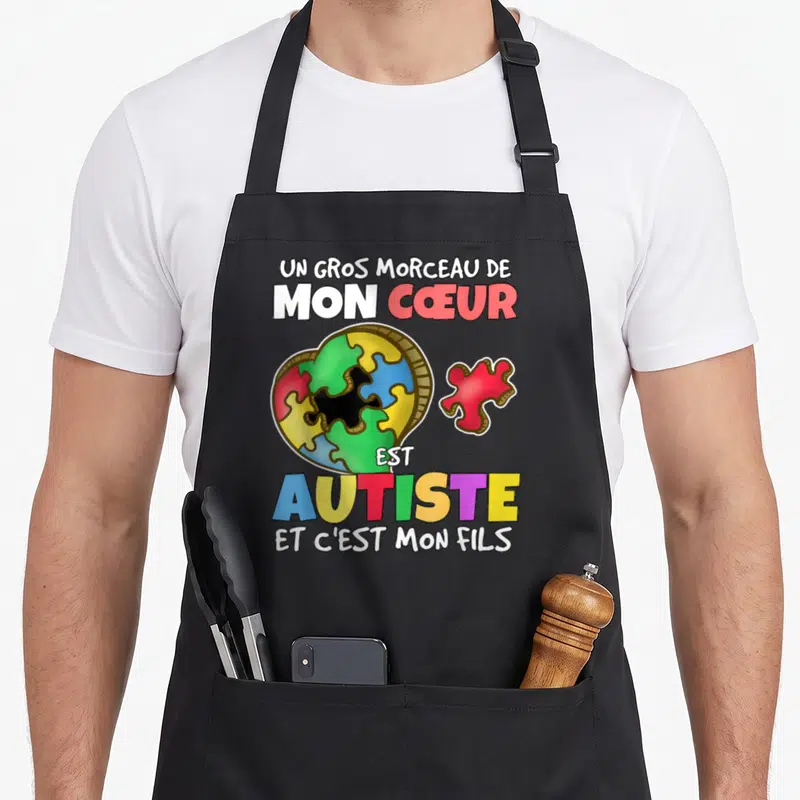 Tablier de cuisine noir avec un cœur en puzzle multicolore et un texte affirmant l'autisme du fils imprimé en lettres vives.