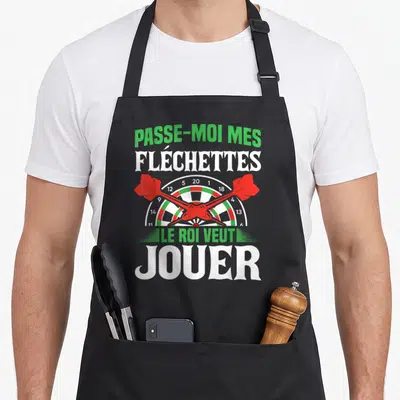 Tablier de cuisine noir avec une cible de fléchettes, trois fléchettes rouges et le texte Passe-moi mes fléchettes le roi veut jouer.