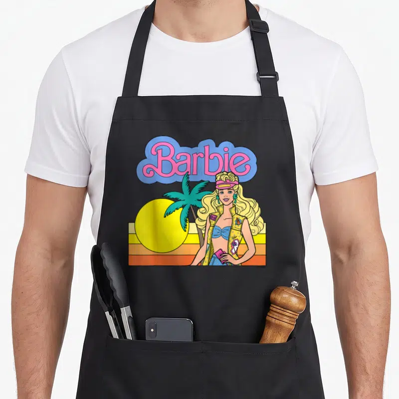 Tablier de cuisine noir avec illustration colorée Barbie, palmier et soleil jaune sur fond rayé orange et jaune.