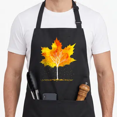 Tablier de cuisine noir avec une illustration de feuille d'érable orange et jaune contenant un arbre blanc aux branches dénudées et des éclats dorés en bas.