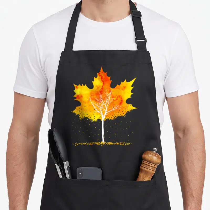 Tablier de cuisine noir avec une illustration de feuille d'érable orange et jaune contenant un arbre blanc aux branches dénudées et des éclats dorés en bas.
