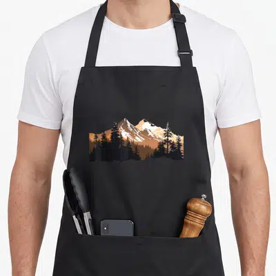 Tablier de cuisine noir avec une illustration de montagnes alpines enneigées dominant une forêt d'épicéas sombres sur fond orange.