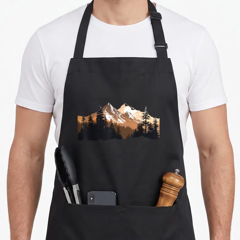 Tablier de cuisine noir avec une illustration de montagnes alpines enneigées dominant une forêt d'épicéas sombres sur fond orange.