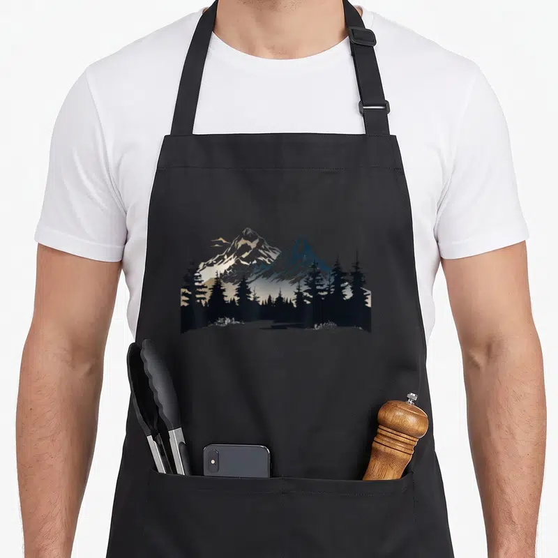 Tablier de cuisine noir avec illustration de montagnes enneigées et silhouettes d'arbres conifères noirs sur fond dégradé.
