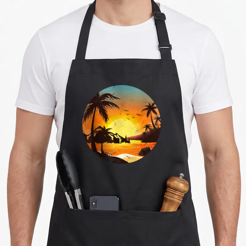 Tablier de cuisine noir avec illustration circulaire de palmiers et montagne au coucher du soleil orange et jaune.
