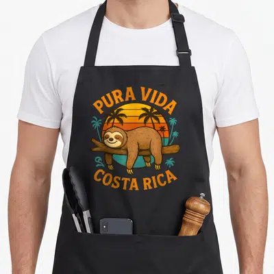 Tablier de cuisine noir avec un paresseux souriant sur une branche devant un soleil orange et des palmiers, encadré par le texte Pura Vida Costa Rica en orange.