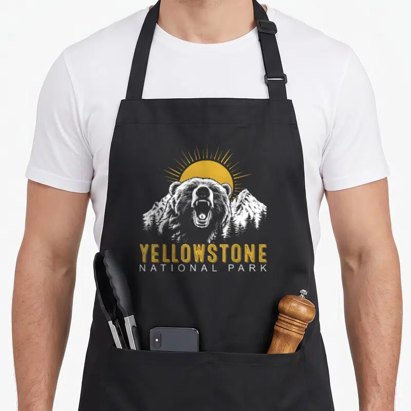 Tablier de cuisine noir avec illustration d'ours rugissant, soleil jaune et texte Yellowstone National Park en style vintage.