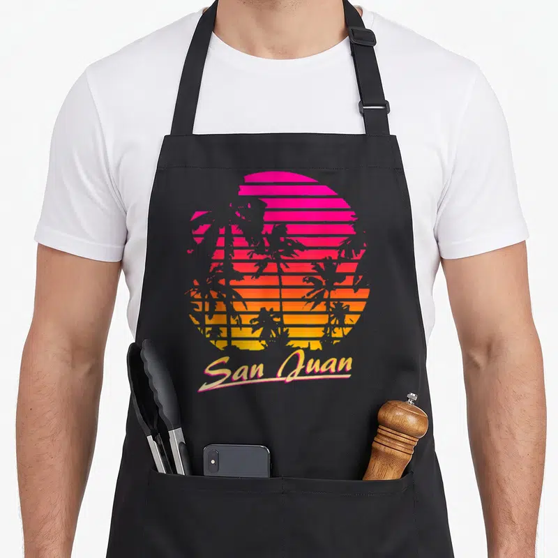 Tablier de cuisine noir avec illustration rétro d'un coucher de soleil rose et orange derrière des palmiers et le texte San Juan.