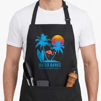 Tablier de cuisine noir avec un flamingo rose, des palmiers bleus et un coucher de soleil rétro sur le devant.