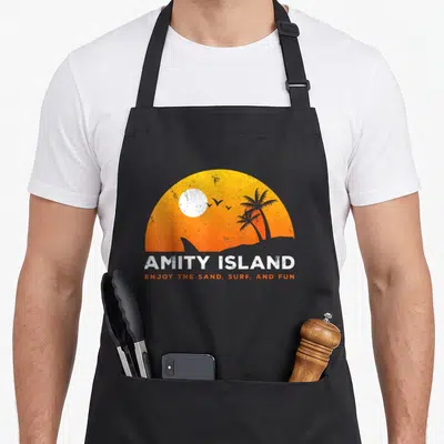 Tablier de cuisine noir avec illustration orange Amity Island sand fun palmiers et vague en silhouette.