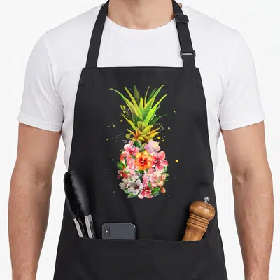 Tablier de cuisine noir avec un ananas composé de fleurs colorées et de feuilles vertes.