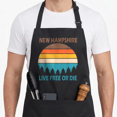 Tablier de cuisine noir avec illustration rétro d'un soleil orange et d'arbres bleus sous le texte New Hampshire Live Free or Die.
