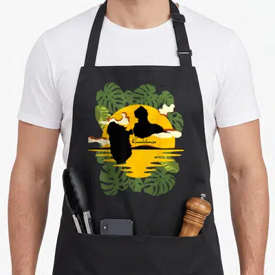 Tablier de cuisine noir avec illustration de coucher de soleil, carte de Guadeloupe et feuilles de monstera vertes.