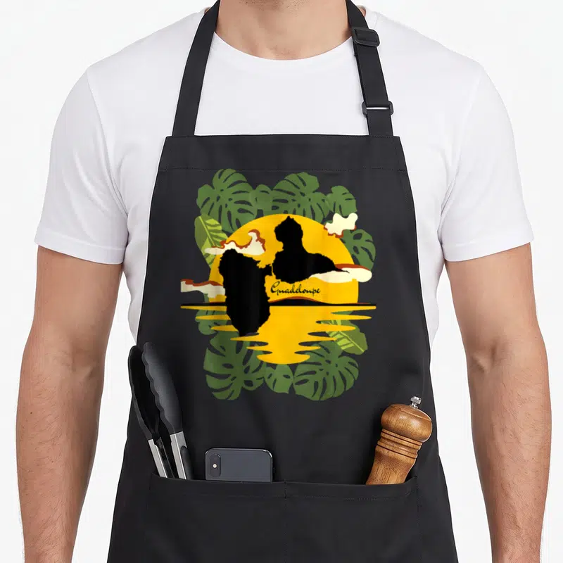 Tablier de cuisine noir avec illustration de coucher de soleil, carte de Guadeloupe et feuilles de monstera vertes.