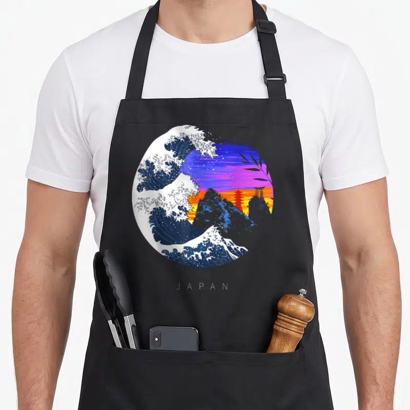 Tablier de cuisine noir avec une grande vague bleue, des montagnes et un temple torii rouge sur fond de coucher de soleil violet et orange.