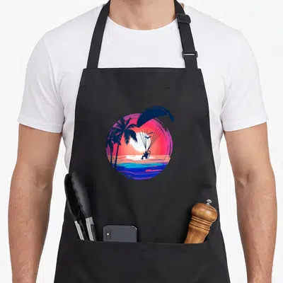 Tablier de cuisine noir avec illustration circulaire d'un paramoteur, palmiers et coucher de soleil rose sur l'eau.